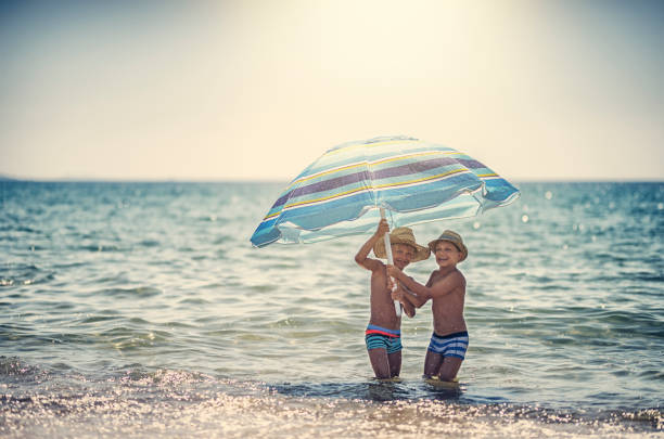 Happy laughing kids are playing on a beach on a sunny summer day. Kids are standing in sea and holding a big beach umbrella.
