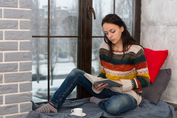 istock Young attractive woman sitting on the windowsill and reading a book 871595120