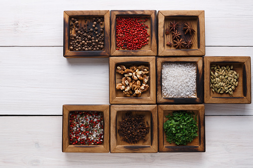 Spices assortment on white rustic background. Wooden boxes with flavorings making frame. Salt, peppers, coriander seeds, cloves, walnuts and green parsley, copy space