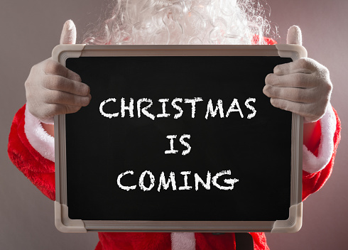 Santa Claus holding a black chalk board written with CHRISTMAS IS COMING