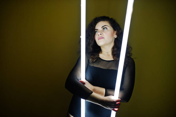 istock Curly brunette plus size model with two long led lamp against yellow wall. 871634486