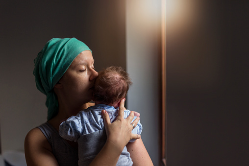 Caucasian Pensive Woman in headscarf, fighting breast cancer while holding her newborn baby relaxing in cancer treatment hospital, patient standing next to hospital window. Mother and baby son. Sleepy little child with mom, eyes closed.