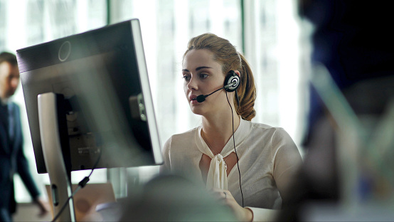 A good looking woman sitting at a desk, surrounded by her co-workers, communicating with someone using a headset & referring to her computer display while she talks on the phone.