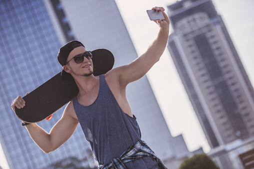 Stylish guy in casual clothes, cap and glasses is holding a skateboard and smiling while doing selfie using a smart phone