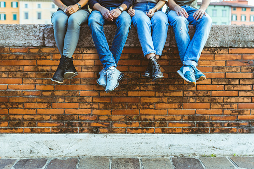 Legs of four teenagers sitting on a bricks wall. Close up view on bottom part of the body, two girls and two boys sitting together in the city. Friendship and lifestyle concepts