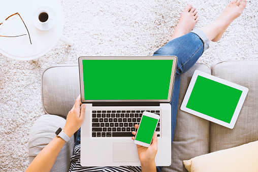 Woman sitting legs crossed on a couch, using green screen laptop, smart phone, digital tablet. Using green screen wireless device at home.