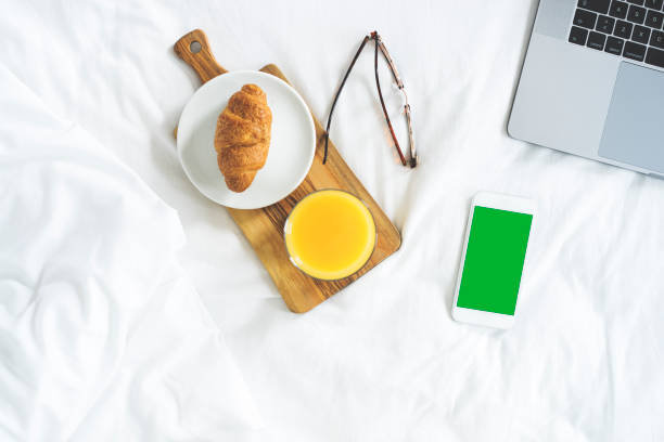 Green screen smart phone on bed in the morning. Orange juice, croissant and glasses in front of laptop.