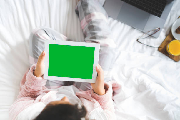 Young woman wearing pyjamas sitting cross legged in bed. Using green screen digital tablet at home.
