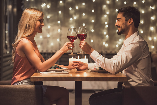 Beautiful young couple is looking at each other, clinking glasses of wine together and smiling during their date in a restaurant