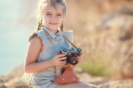 A little girl of 7 years with two braids, one sits outdoors on a Sunny summer day, on top of a cliff against the sea with an old retro suitcase,dressed in a light blue jumpsuit,holding hands an old camera,takes photos of nature