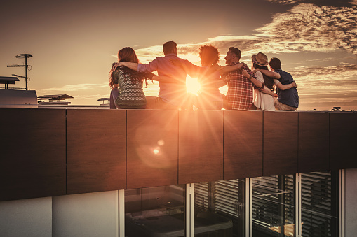 Back view of large group of friends sitting embraced on a roof and enjoying in sunset.