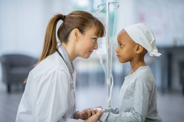 Doctor And Child A female Caucasian doctor and a young girl of African descent are indoors in a hospital room. The girl has cancer, and she is wearing a hat. She is being comforted by her doctor while being hooked up to an IV. hospital depression sadness bed stock pictures, royalty-free photos & images
