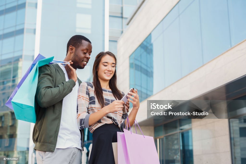 Multiethnic couple shopping together Multiethnic happy couple reading message in smartphone after shopping together, walking with bags at mall, copy space Retail Stock Photo