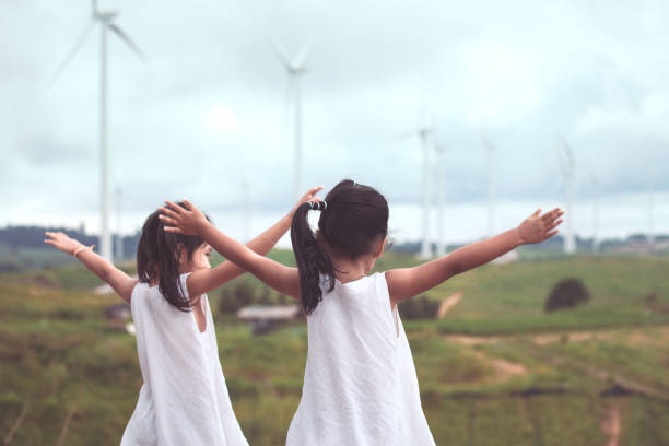 rückansicht der beiden asiatischen kind mädchen heben ihre arme blick auf wind-turbine-feld - fuel and power generation power wind power supply stock-fotos und bilder