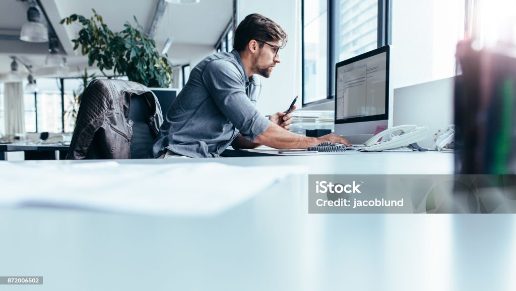 Young businessman in office working on computer Young man sitting in office and working on desktop pc. Businessman looking at computer monitor while working in office. Office Stock Photo