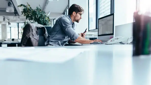 Young man sitting in office and working on desktop pc. Businessman looking at computer monitor while working in office.