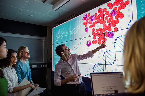 Group of business professionals in a dark room standing in front of a large data display screen with information.