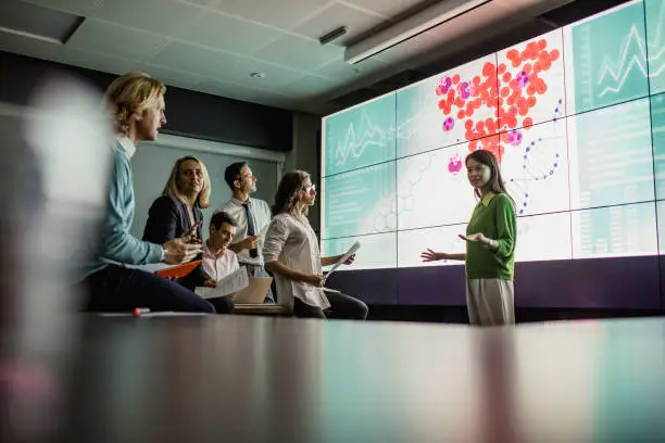 Group of business professionals in a dark room standing in front of a large data display screen with information.