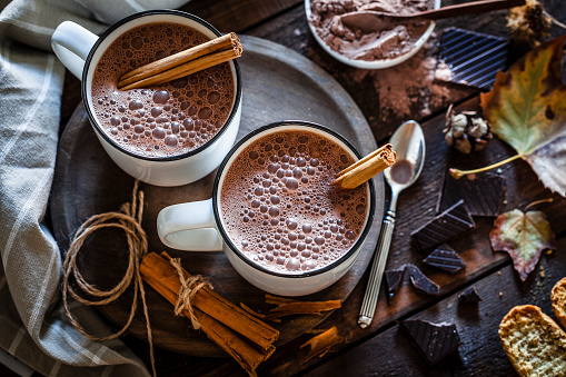 Two hot chocolate mugs shot from above on rustic wooden table. Some cinnamon sticks are on the table and inside the mugs and chocolate pieces are beside the mugs placed directly on the table. Low key DSRL studio photo taken with Canon EOS 5D Mk II and Canon EF 100mm f/2.8L Macro IS USM