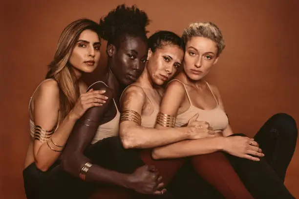 Group of diverse women sitting together against brown background. Multi ethnic females with different skin tones in studio.