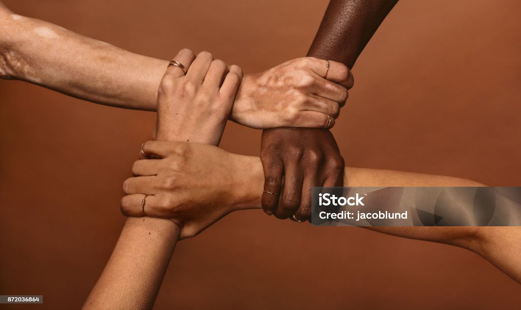 Unity in diversity Four diverse women holding each others wrists in a circle. Top view of female hands linked in the lock against brown background. Multiracial Group Stock Photo