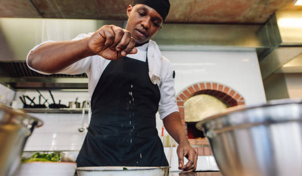 Chef sprinkling spices on dish in commercial kitchen. African male cook preparing food in restaurant kitchen, adding seasoning.