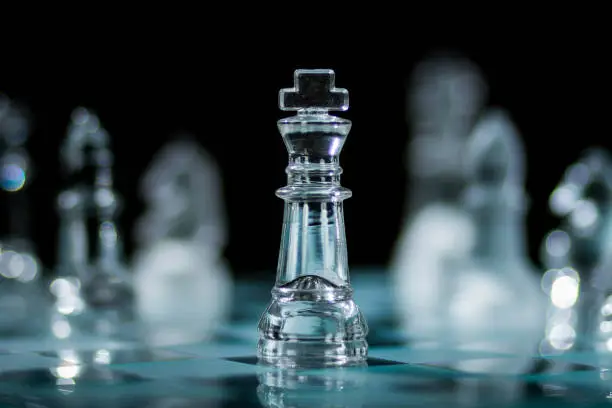 A glass king chess piece standing on a chess board and surrounded by other pieces out of focus of both colours