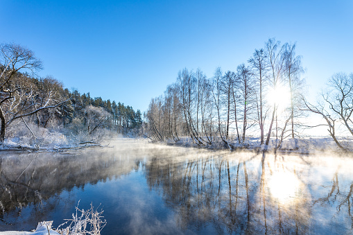 Steam over the winter river with snow on riverside. Clear blue sky background.