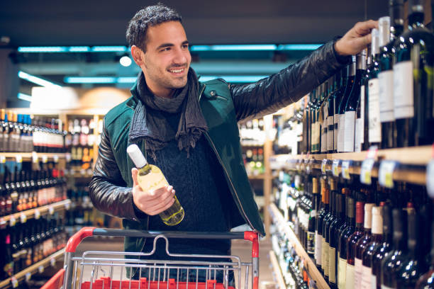 Young man in a supermarket choosing wine Young man with a shopping cart is in a supermarket picking up wine. alcohol shop stock pictures, royalty-free photos & images