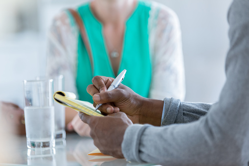 In this closeup, two unrecognizable people, a man and a woman, sit at a table containing two half full glasses of water.  The woman has a handbag on her shoulder and the man writes on a yellow notebook with a pen.