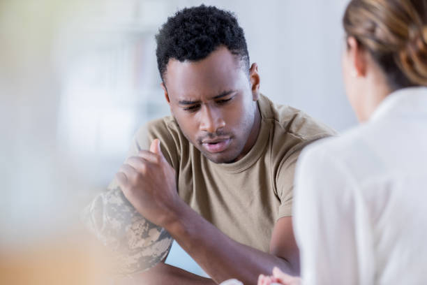 Distraught military veteran talks with counselor In this closeup, a young man wearing a military issue t-shirt and holding a camouflage cap looks down with a pained expression.  He is sharing his problems with an unrecognizable counselor. black military man stock pictures, royalty-free photos & images