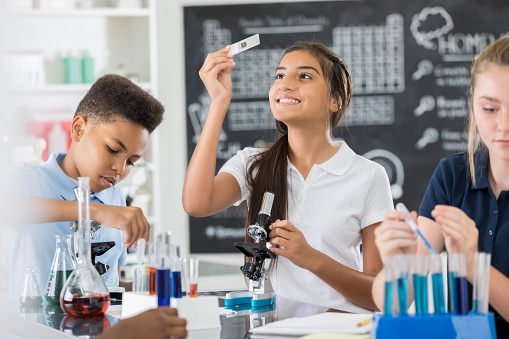 Intelligent Hispanic middle school girl examines a microscope slide during science lap. Other students are working beside her.