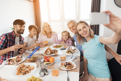 A woman in a blue T-shirt is sitting at a festive table and making a selfie on a white smartphone. Her relatives are posing behind her. Everyone is celebrating Thanksgiving Day