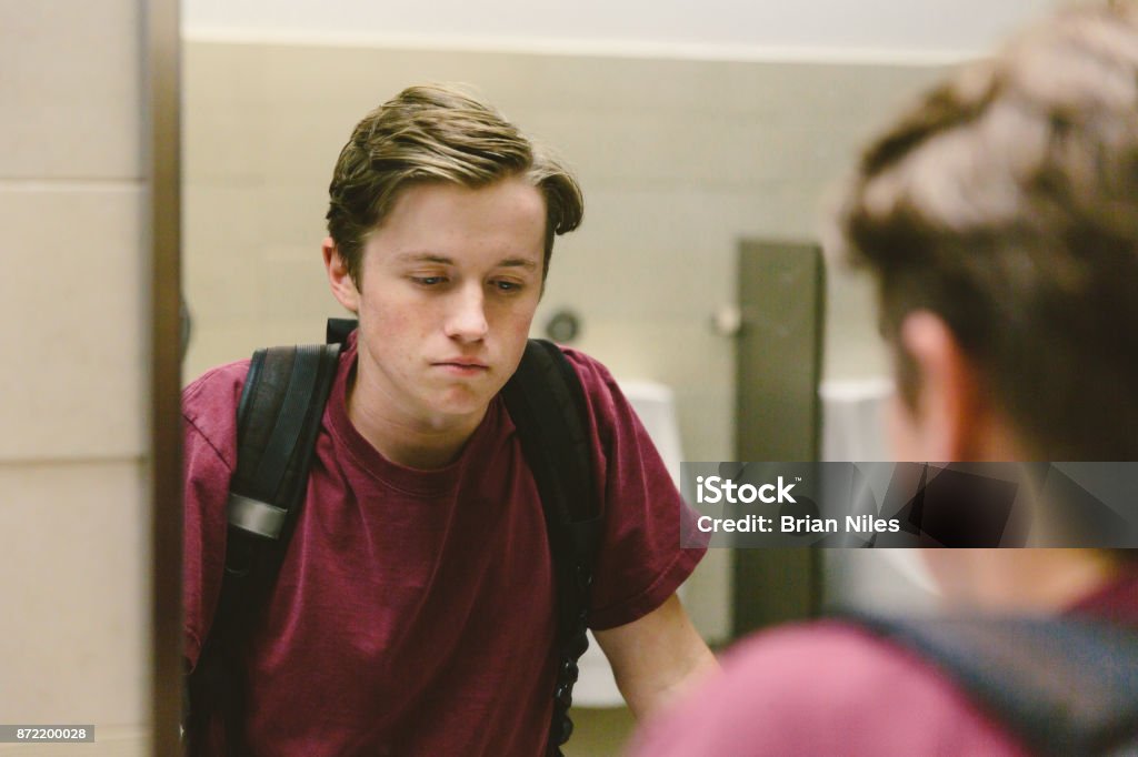 Depressed teen looks at himself in bathroom mirror Depressed teen student helplessly stares at his reflection in bathroom mirror. Teenager Stock Photo