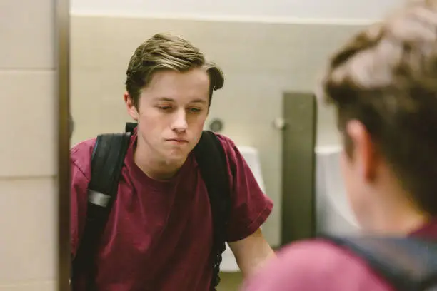 Depressed teen student helplessly stares at his reflection in bathroom mirror.