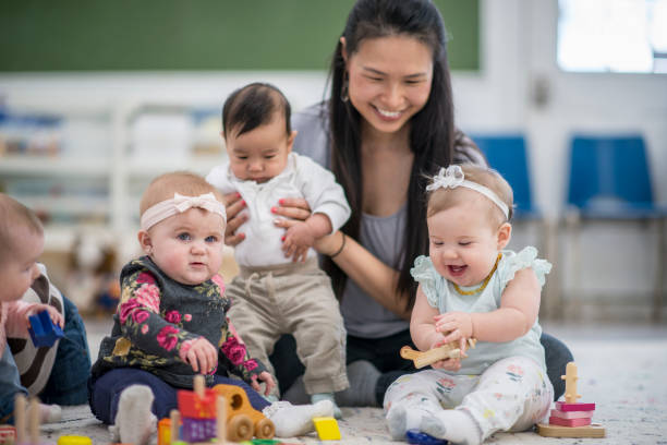 Happy Babies A multi-ethnic group of babies are indoors in a daycare center. They are wearing casual clothing. They are playing with toys along with their babysitter. One baby is laughing while playing with a wooden block. asian nanny stock pictures, royalty-free photos & images