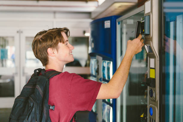 Teen boy paying with mobile phone at vending machine Student teenage boy wearing backpack uses mobile phone to pay for snack and drink at vending machine vending machine stock pictures, royalty-free photos & images