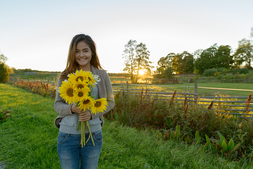 Young happy Asian woman holding sunflowers behind peaceful scenery of grass field