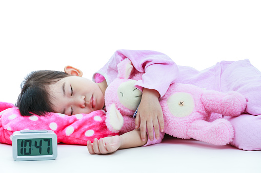 Healthy children concept. Asian child with doll sleeping peacefully. Adorable girl in pink pajamas taking a nap with alarm clock in foreground. On white background.