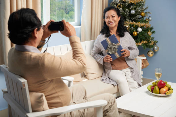 Photographing with present Senior man taking photo of his wife near Christmas tree vietnamese old man portrait stock pictures, royalty-free photos & images