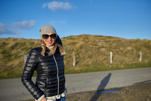 Trendy woman wearing a leather jacket, woollen cap and sunglasses standing on a rural road in evening light with copy space smiling at the camera
