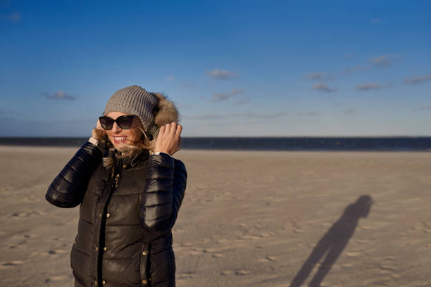 Trendy woman in a warm autumn outfit with sunglasses standing on a beach at sunset casting a long shadow across the sand looking to the side with a happy smile