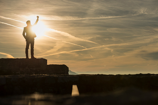 Triumphant businessman greeting a new day standing outdoors on a wall with his fist raised against a sun with copy space.