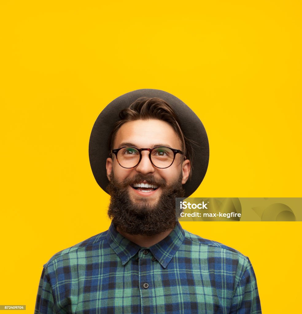 Content young man on orange Portrait of young bearded hipster in hat and eyeglasses looking happily up on orange background. Men Stock Photo