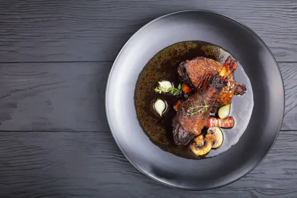 portion of tasty chicken stew - thigh and drumstick braised with wine, herbs, mushrooms and vegetables served on black plate on wooden table, festive Burgundian dish - Coq Au Vin, view from above