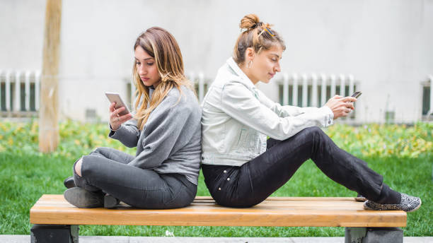 istock Two girl friends using cellphones on park bench 872484148