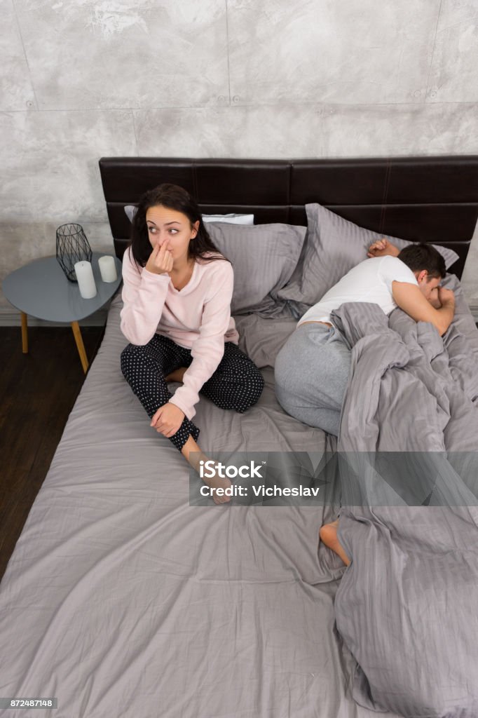 Young woman covers her nose because of her boyfriend, who farted while sleeping in the bed Young woman covers her nose because of her boyfriend, who farted while sleeping in the bed, both wearing pajamas in the bedroom in loft style Diarrhea Stock Photo