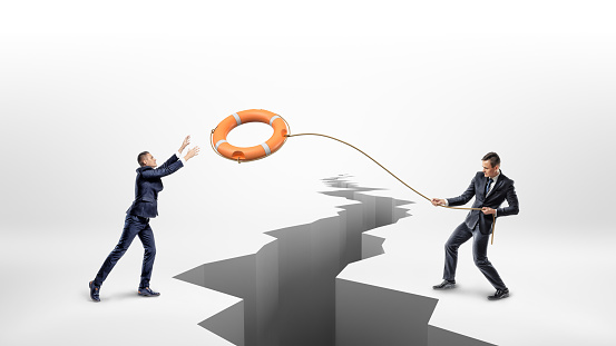 A businessman throws a lifeline with an orange life buoy over a large earthquake crack to another businessman. Business and cooperation. Help your colleagues. Workplace assistance.