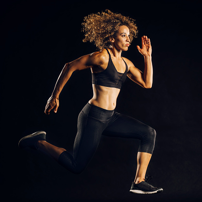 An athletic African American woman running mid-air in fitness clothing, in studio with a black background.