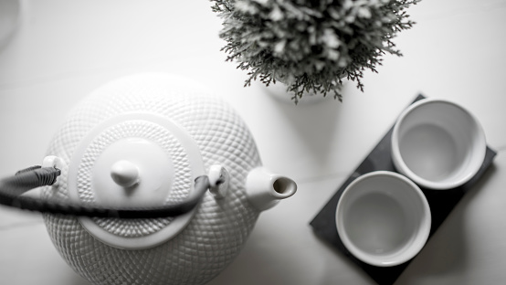 Closeup of breakfast or afternoon tea: a white porcelain cup of hot tea/coffee, a mug of milk, a tray of pastries and a teapot on a hardwood table with a green garden view.  Blurred large, high windows with blinds and a terrace and garden view (chairs, tables, and trees) in the background. A slight vintage effect was added. 3D rendered image.